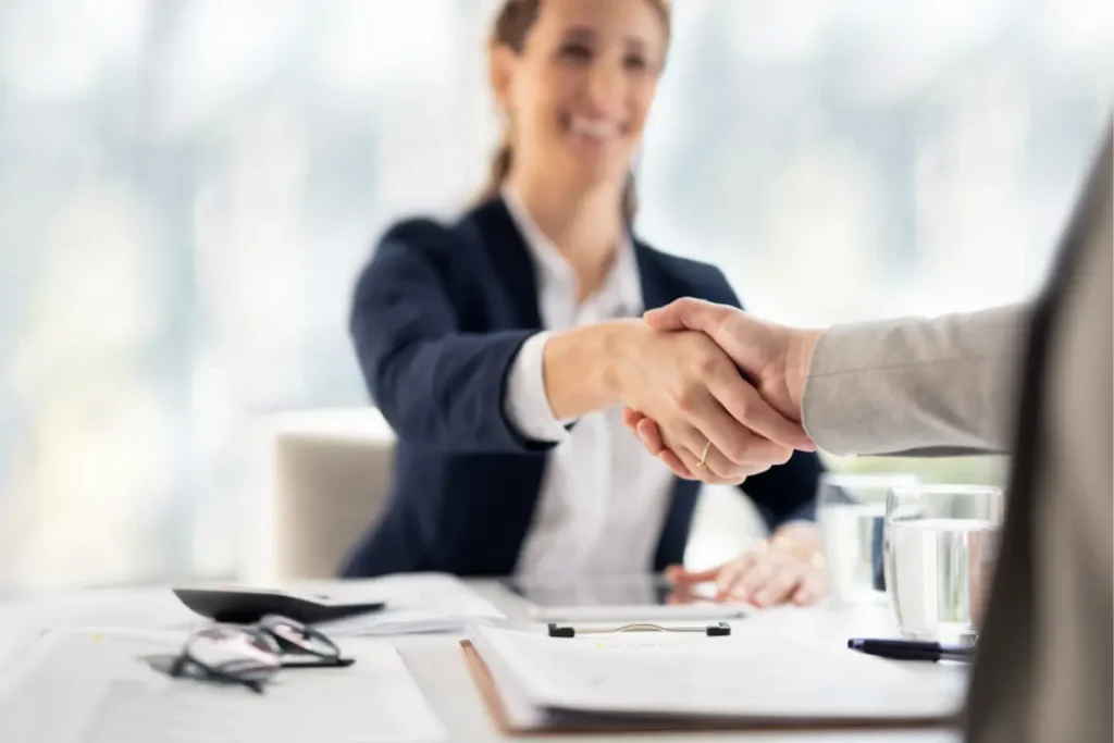 Zoom sur une poignée de main entre deux personnes, symbolisant la conclusion d’un accord. À l’arrière-plan, une femme apparaît floue ; elle est l’une des deux personnes qui se serrent la main. La scène se déroule dans un bureau professionnel.